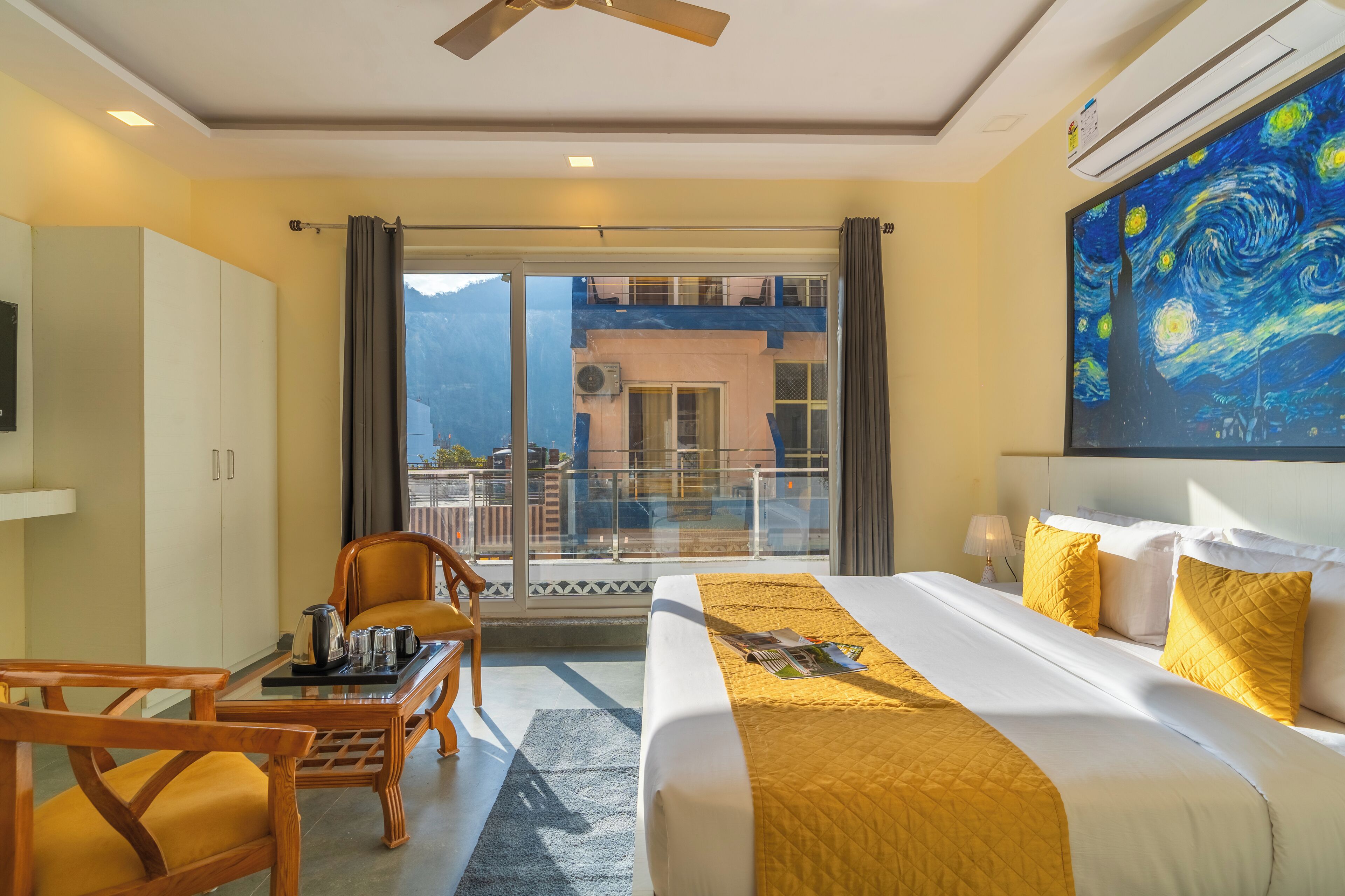 Habitación de hotel con cama, mesa con sillas, balcón con vistas a las montañas y un cuadro en la pared.