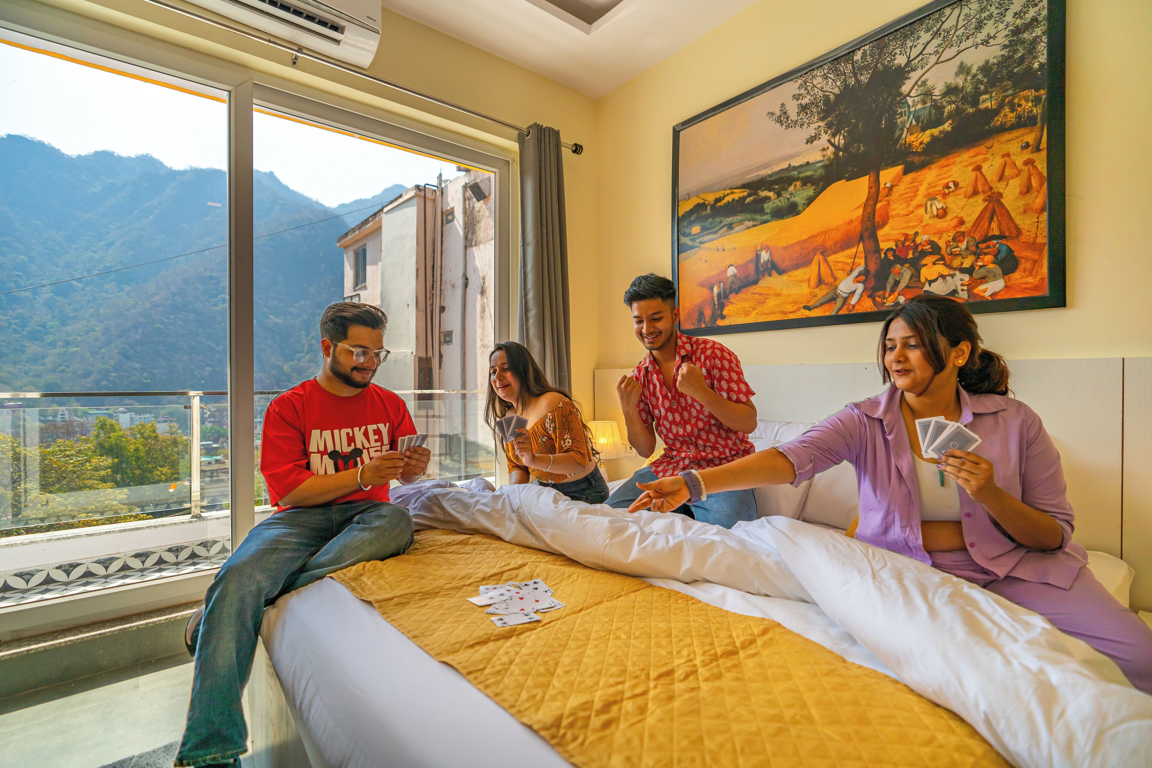 A group of people playing cards on a bed with a mountain view through a large window.