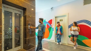 An indoor corridor with a glass elevator, a colorful wall mural, and a tiled floor. - The Social Stays Rishikesh (Narendranagar)