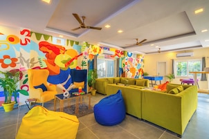 Un salón luminoso con murales coloridos, un sillón amarillo, sofás verdes y un asiento de peluche azul.
