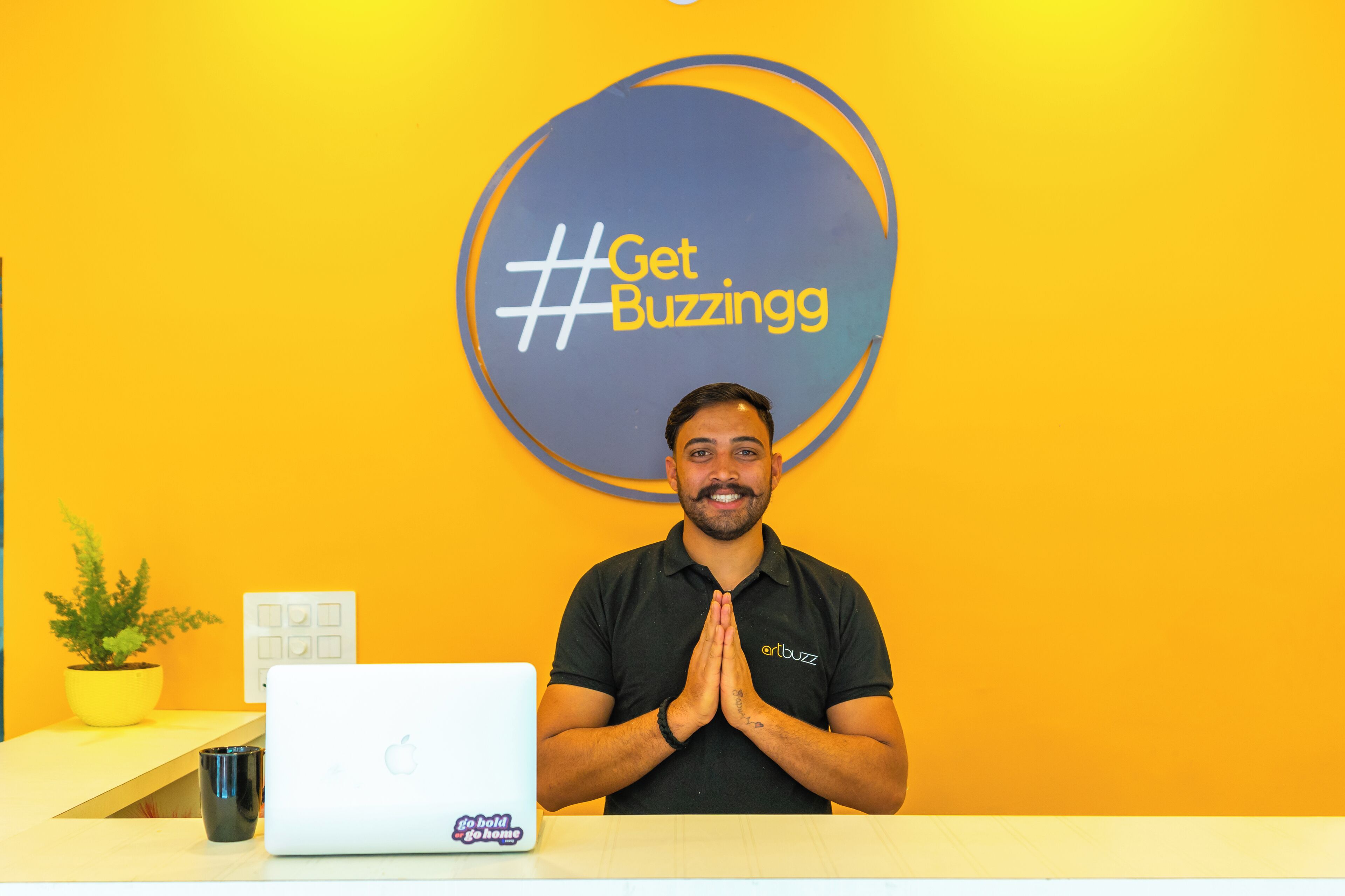 Una persona de pie detrás de un escritorio con una computadora portátil y un letrero que dice '#GetBuzzingg'.
