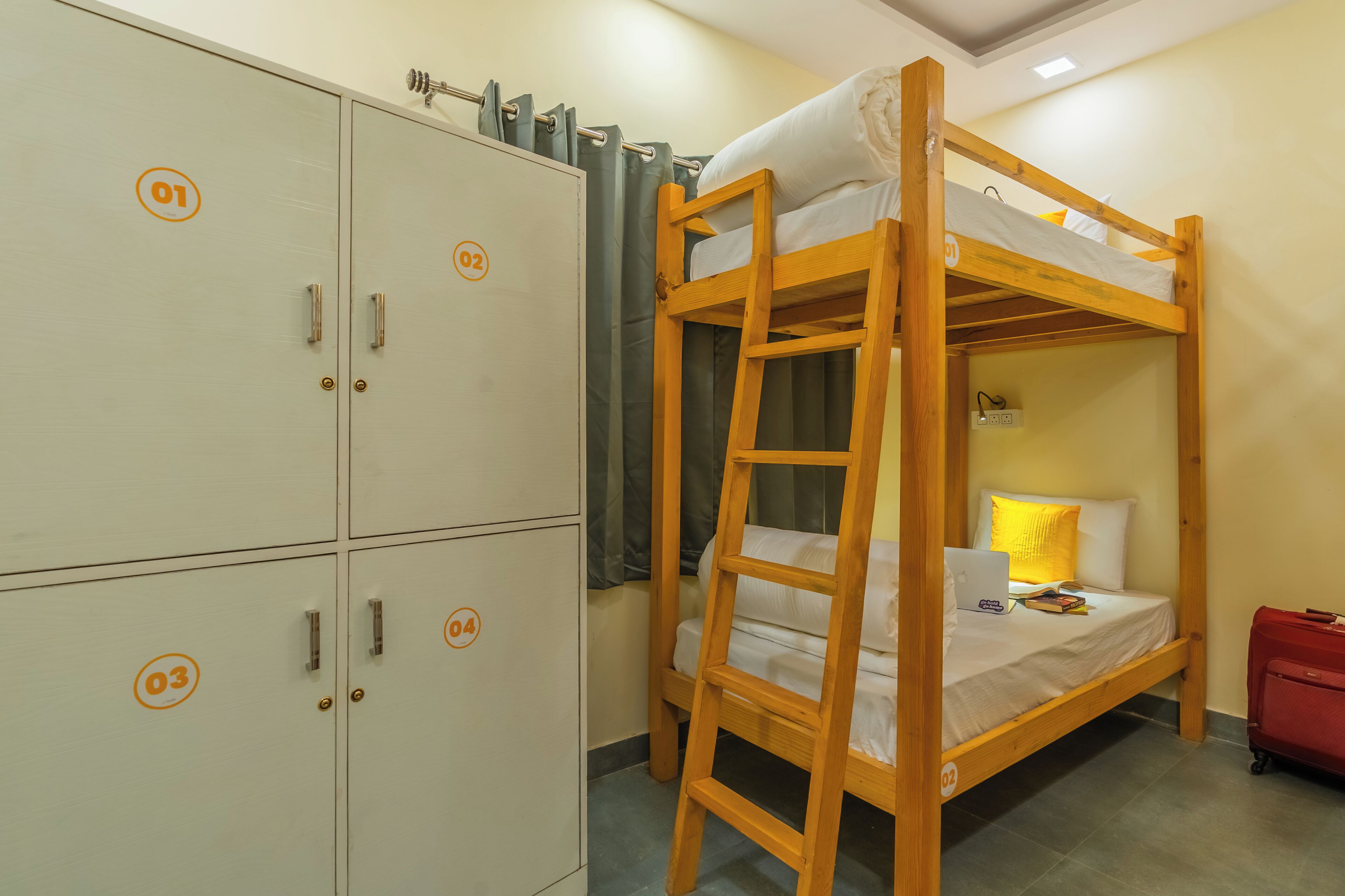 A bunk bed with a laptop and a red suitcase, a wardrobe with numbered doors, and a curtain.