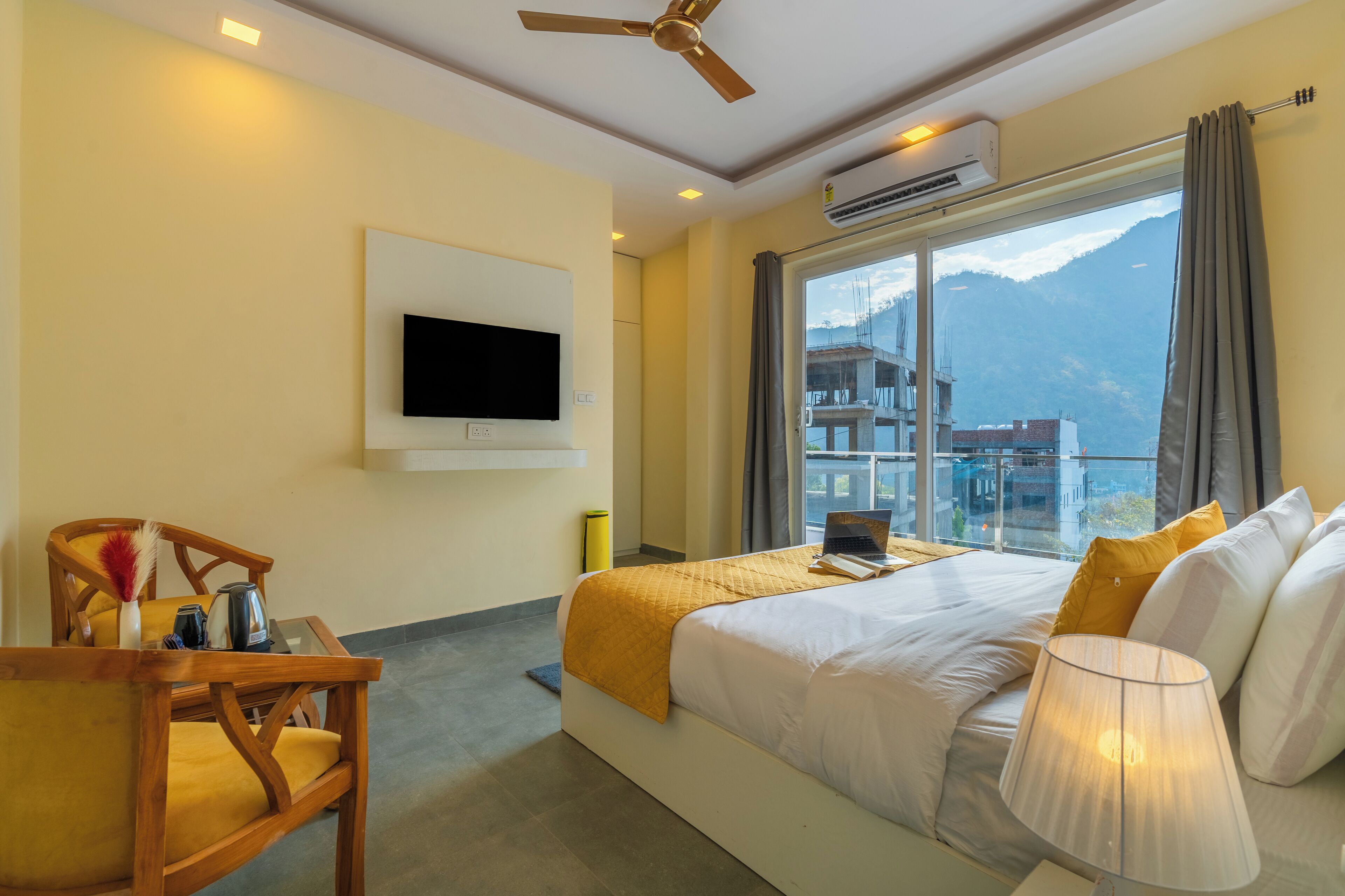 A hotel room with a bed, a desk with a laptop, a chair, a TV mounted on the wall, and a window with a mountain view.
