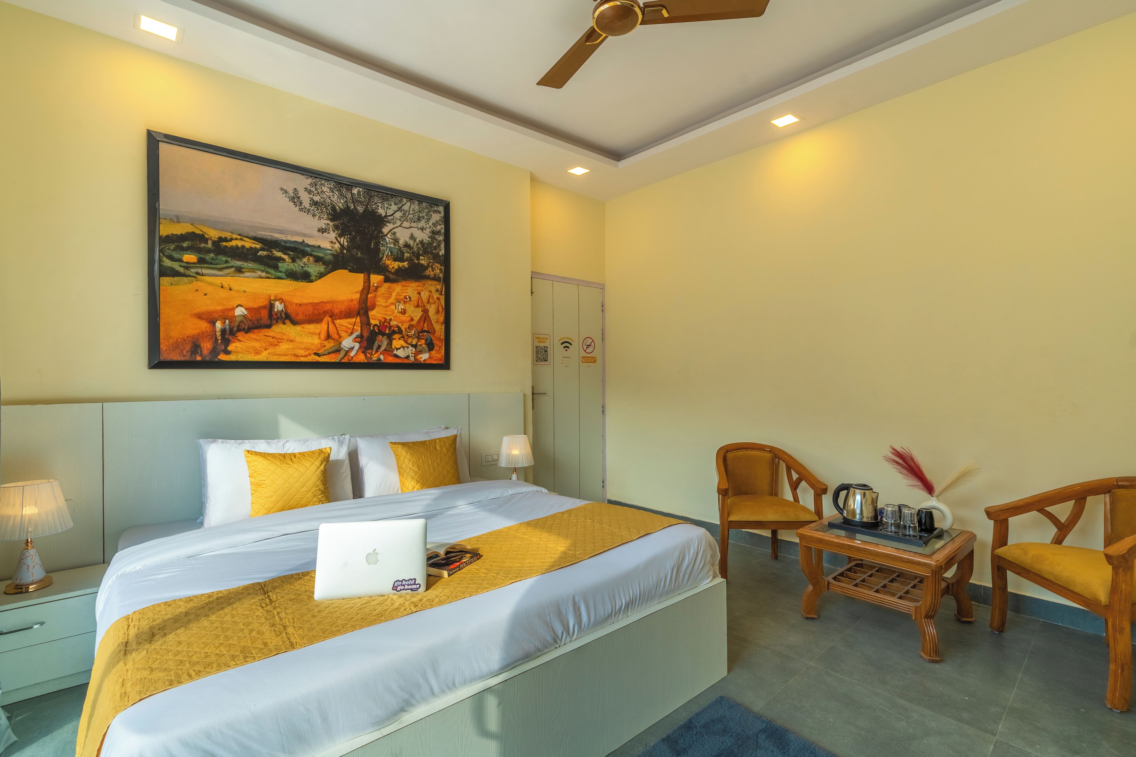 Une chambre d’hôtel avec un grand lit, un bureau avec un ordinateur portable, deux chaises, un tableau au mur et un ventilateur de plafond.