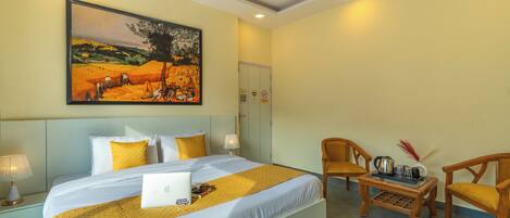 Une chambre d’hôtel avec un grand lit, un bureau avec un ordinateur portable, deux chaises, un tableau au mur et un ventilateur de plafond.