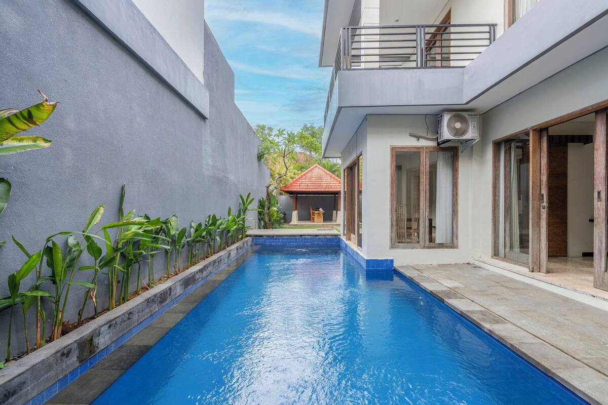 Una piscina con borde de azulejos, rodeada por un muro de hormigón y un edificio con balcón y puertas correderas.