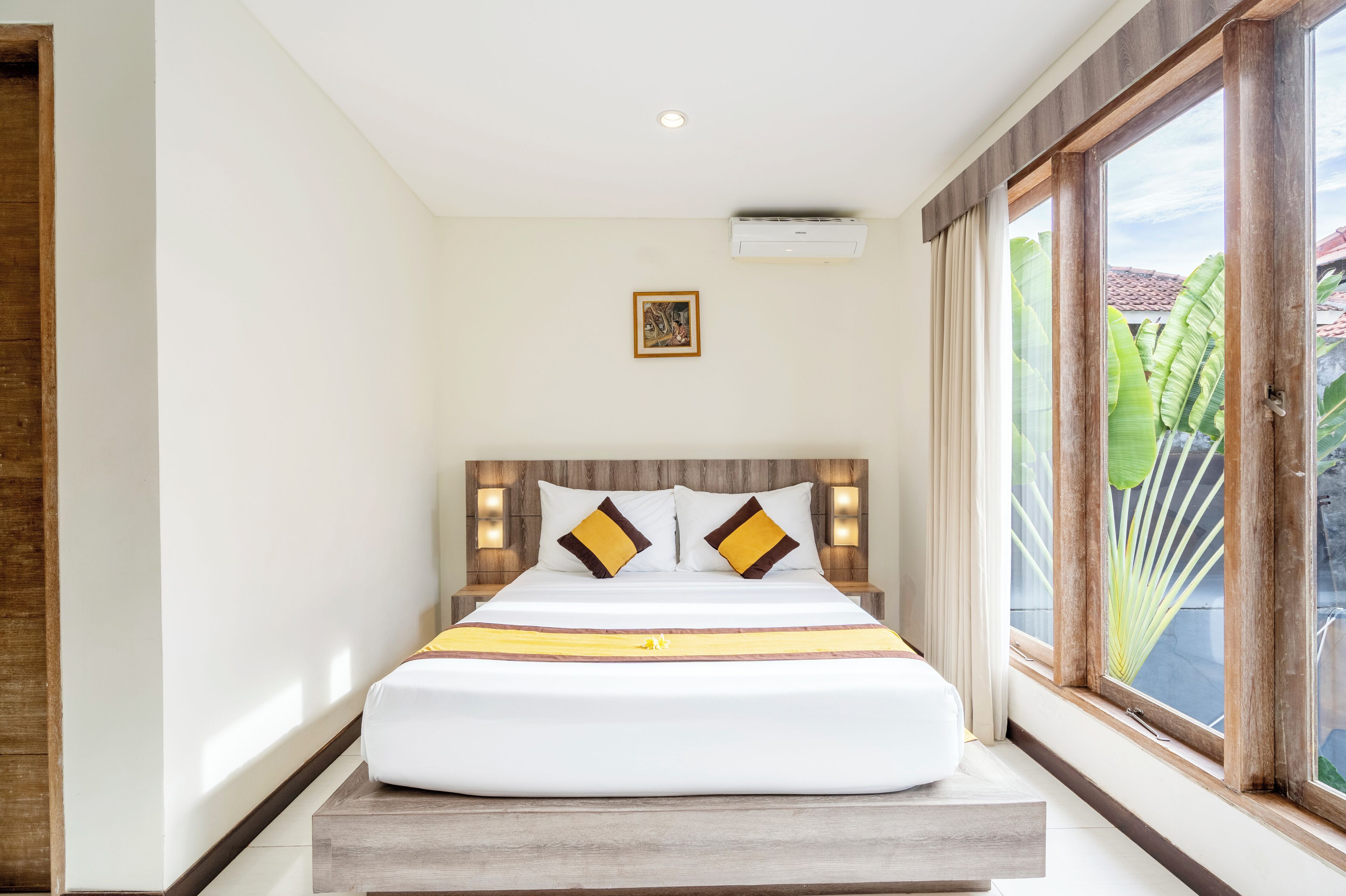 A neatly made bed with a wooden headboard, a small framed picture above it, and a large window with a view of greenery outside.