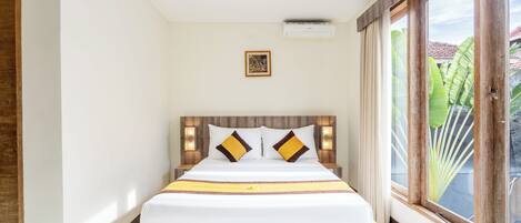 A neatly made bed with a wooden headboard, a small framed picture above it, and a large window with a view of greenery outside.