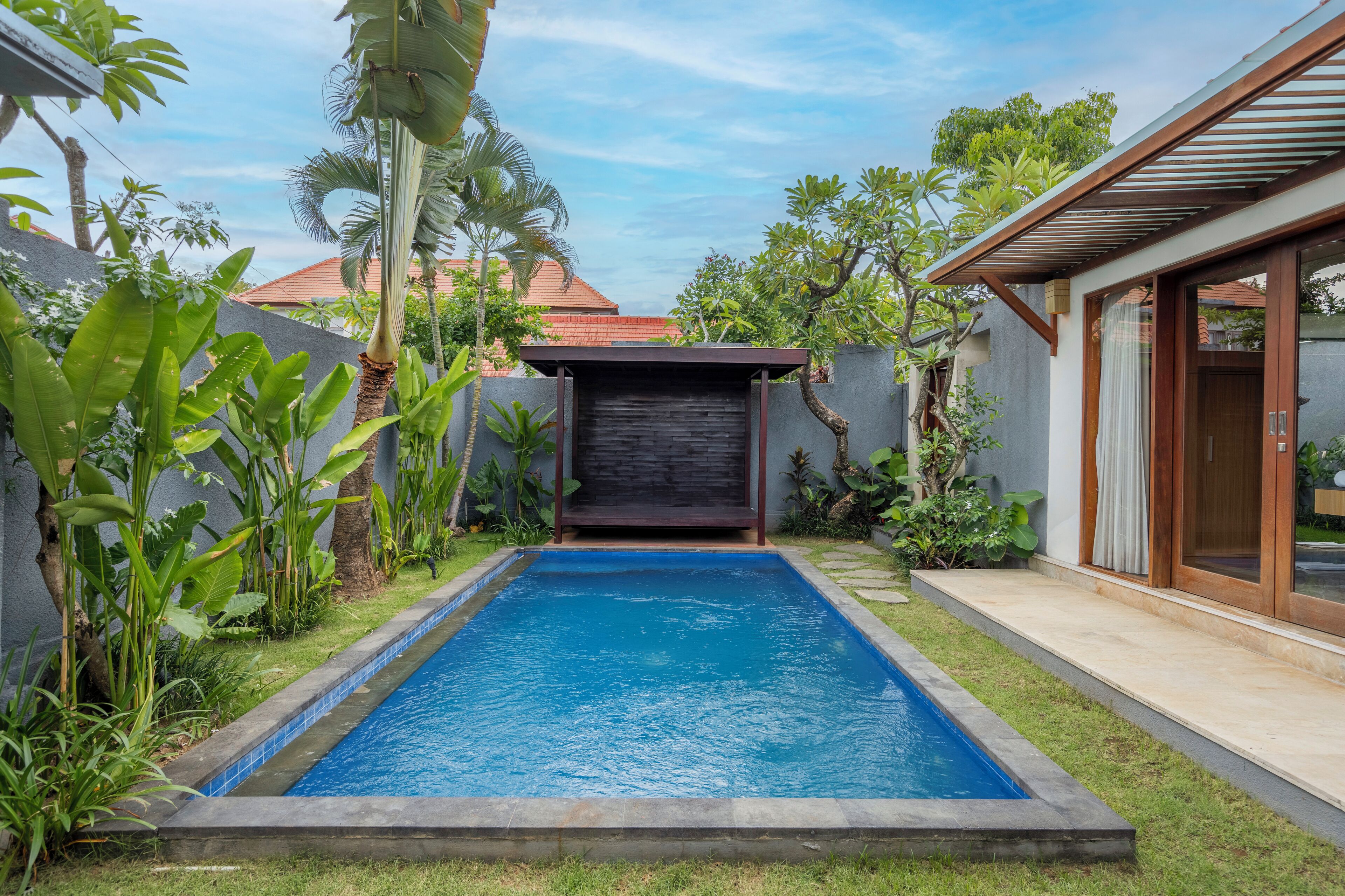 A rectangular swimming pool with a dark cover, surrounded by lush greenery and a small structure with a dark roof.