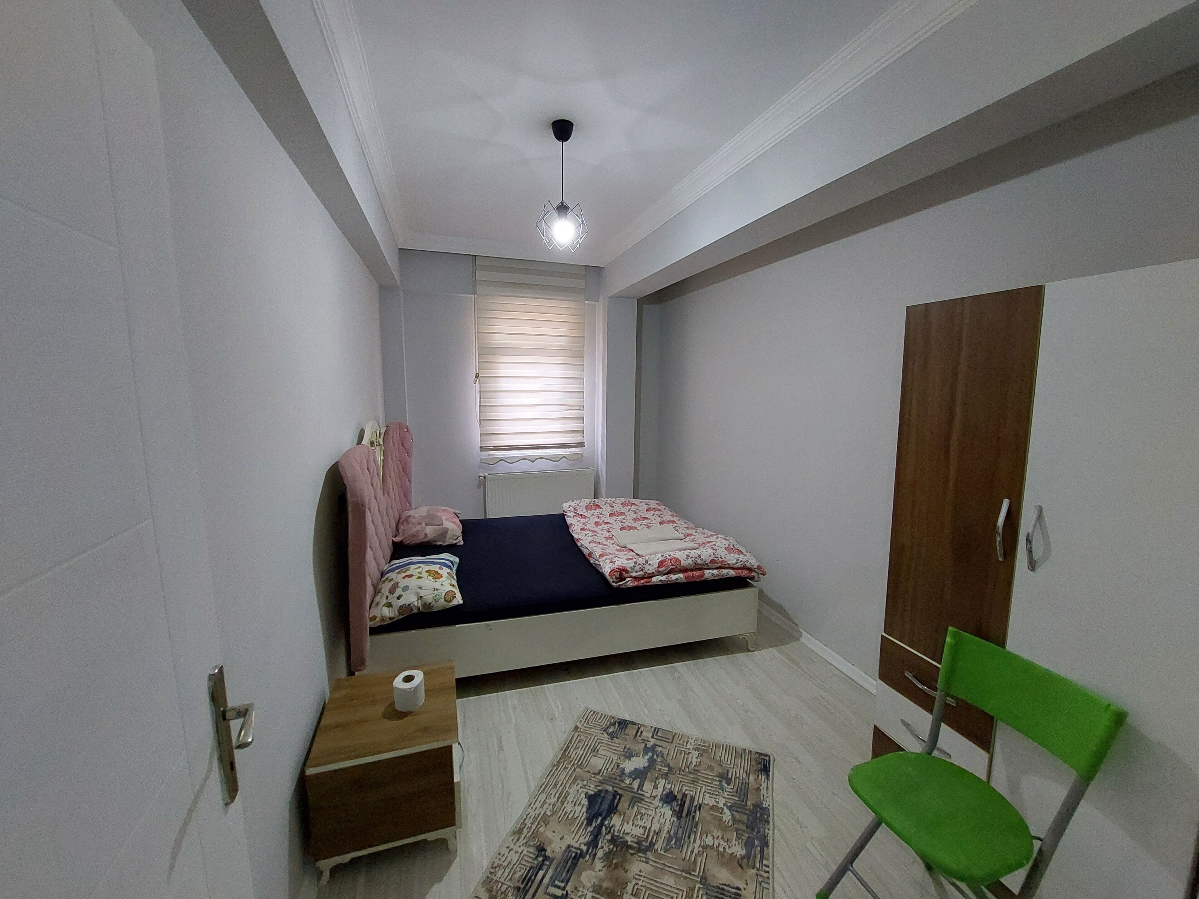 A small, neatly arranged bedroom with a single bed, a wooden nightstand, a green chair, and a wardrobe.