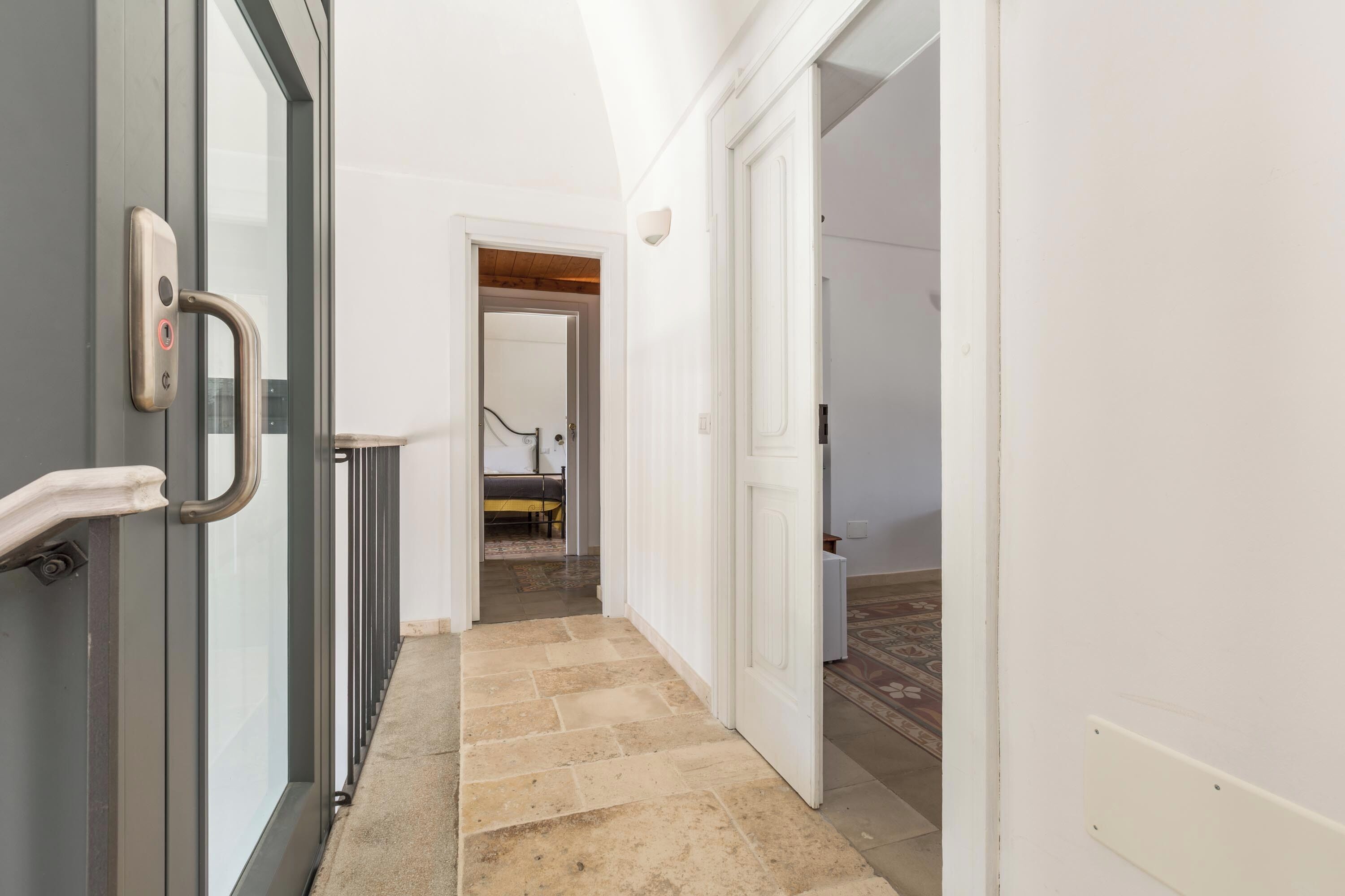 A hallway with a glass door on the left, a white door on the right, and a bed in the background.