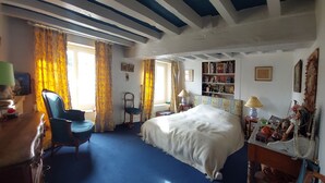 A bedroom with a large bed, a desk with a lamp, a chair, a bookshelf, and a window with curtains.