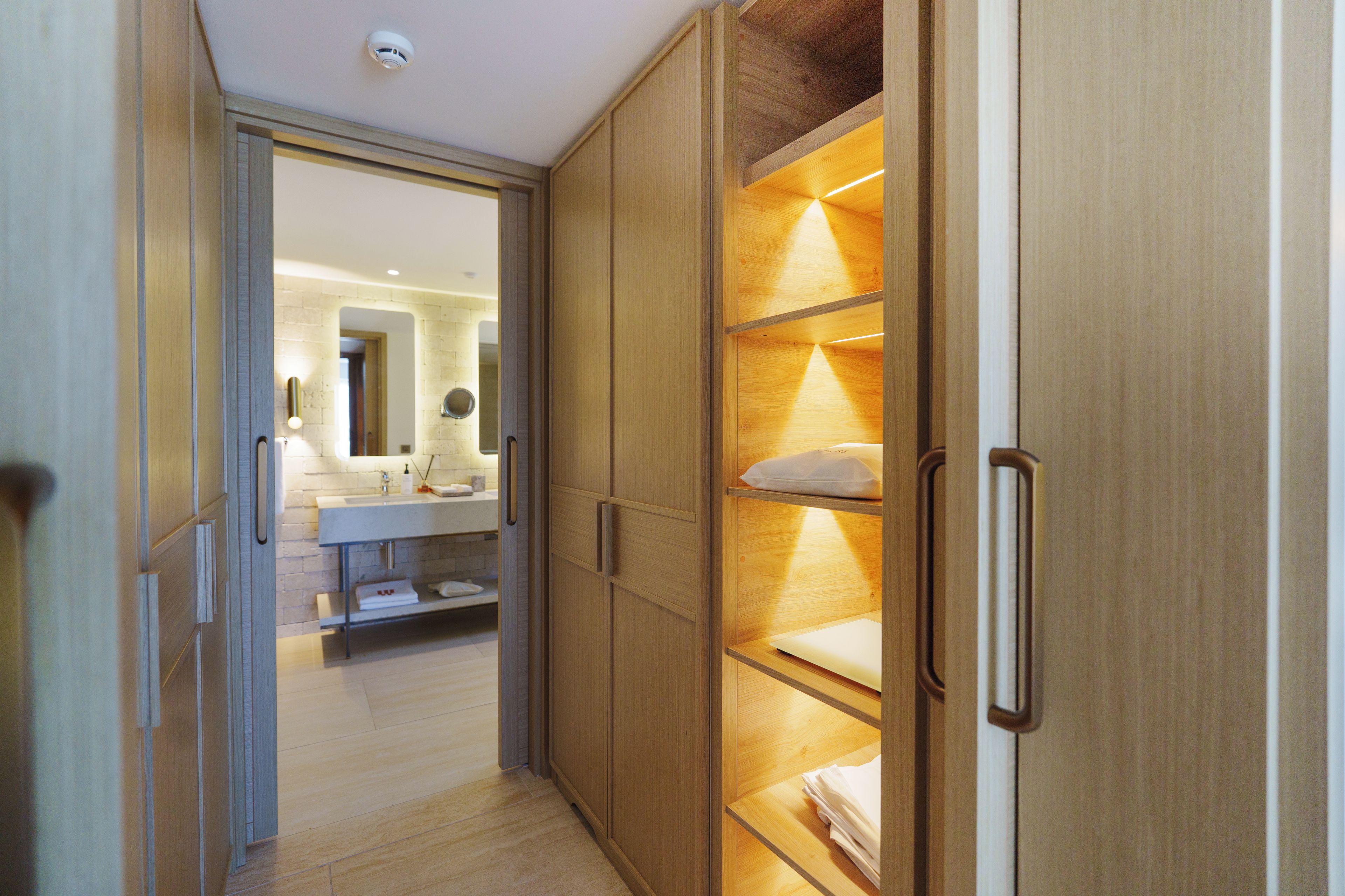 Un dressing avec des portes et des étagères en bois, une salle de bain avec un lavabo et un miroir, et un couloir menant à une autre pièce.