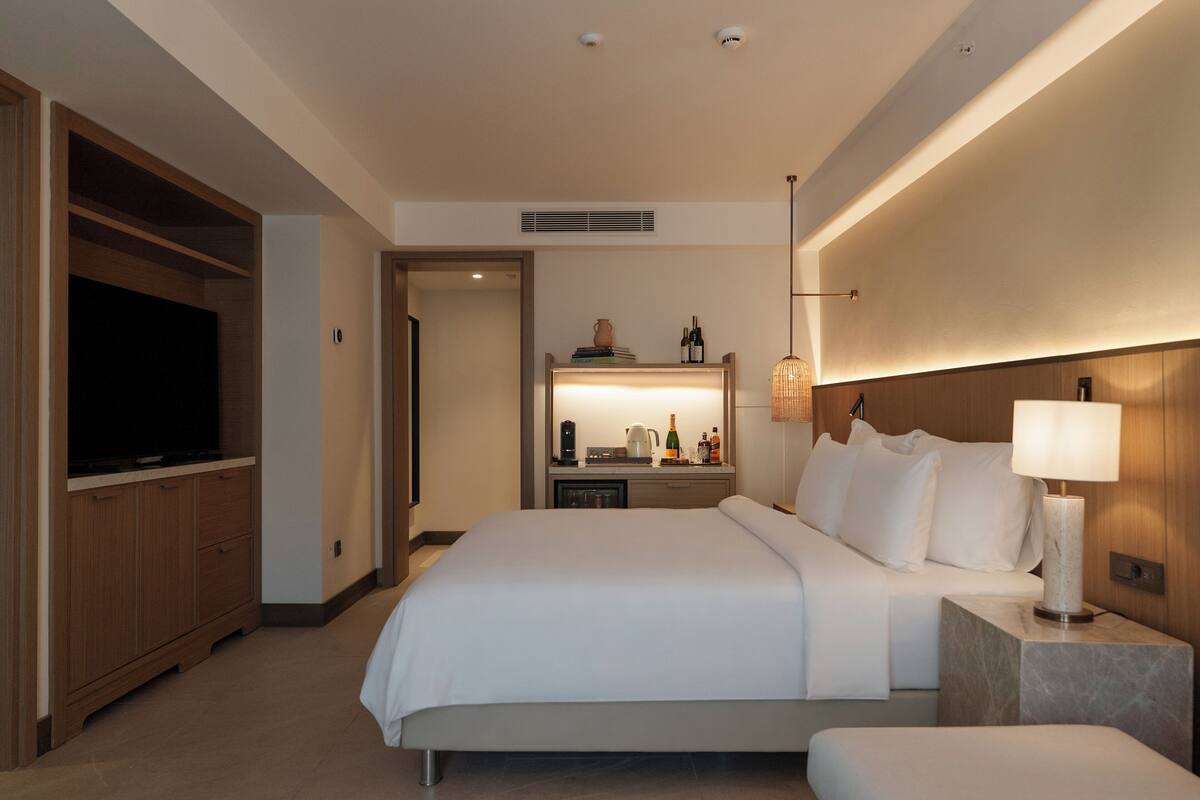 A hotel room with a large bed, a TV, a bedside table with a lamp, and a minibar.