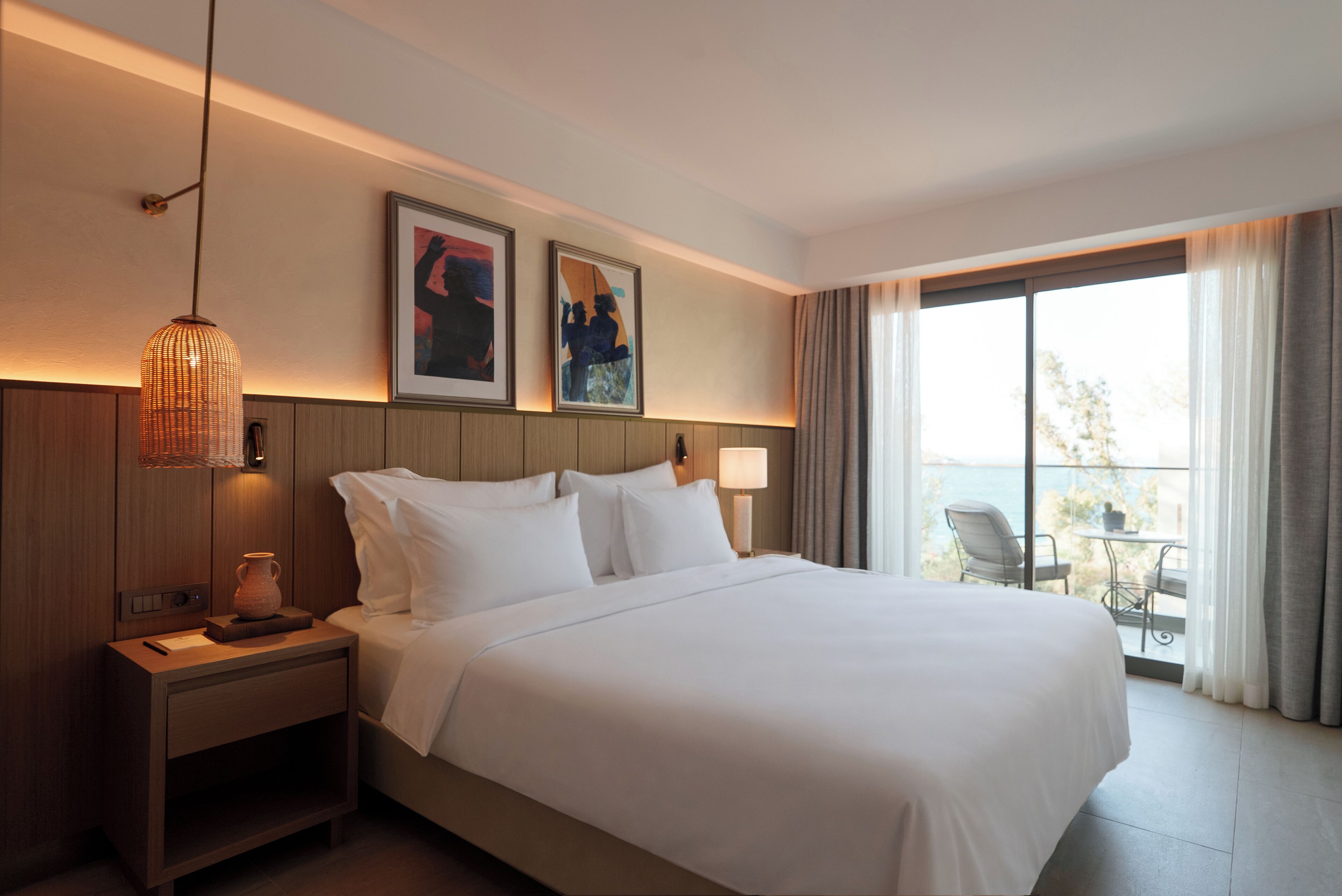 Habitación de hotel con una cama grande, una mesita de noche, una lámpara y vistas al exterior a través de un amplio ventanal.
