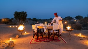 Un décor de dîner en plein désert, avec une table dressée pour deux personnes, entourée de bougies et un chef qui prépare le repas.