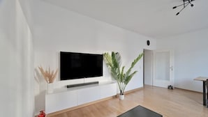 A modern living room with a flat-screen TV mounted on a white wall, a wooden floor, a small black coffee table, and a potted plant. - Goodliving Apartments | Netflix*Office*Parking (Essen)
