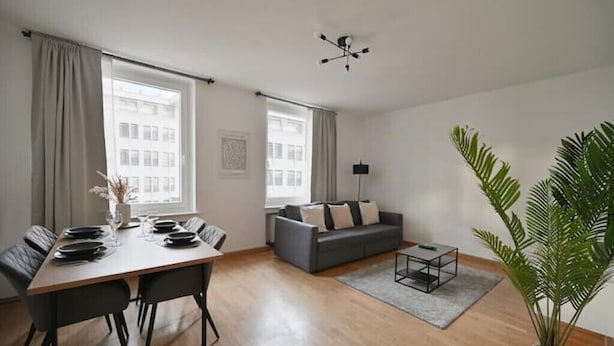 A modern living room with a dining area, a grey sofa, a coffee table, and a large plant. - Goodliving Apartments | Netflix*Office*Parking (Essen)
