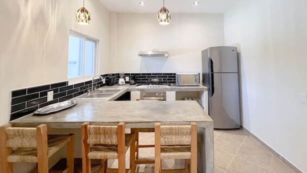 A modern kitchen with a central island, black and white backsplash, stainless steel appliances, and wooden chairs. - Best Location + Clean, Safe, Just Built (Sayulita)