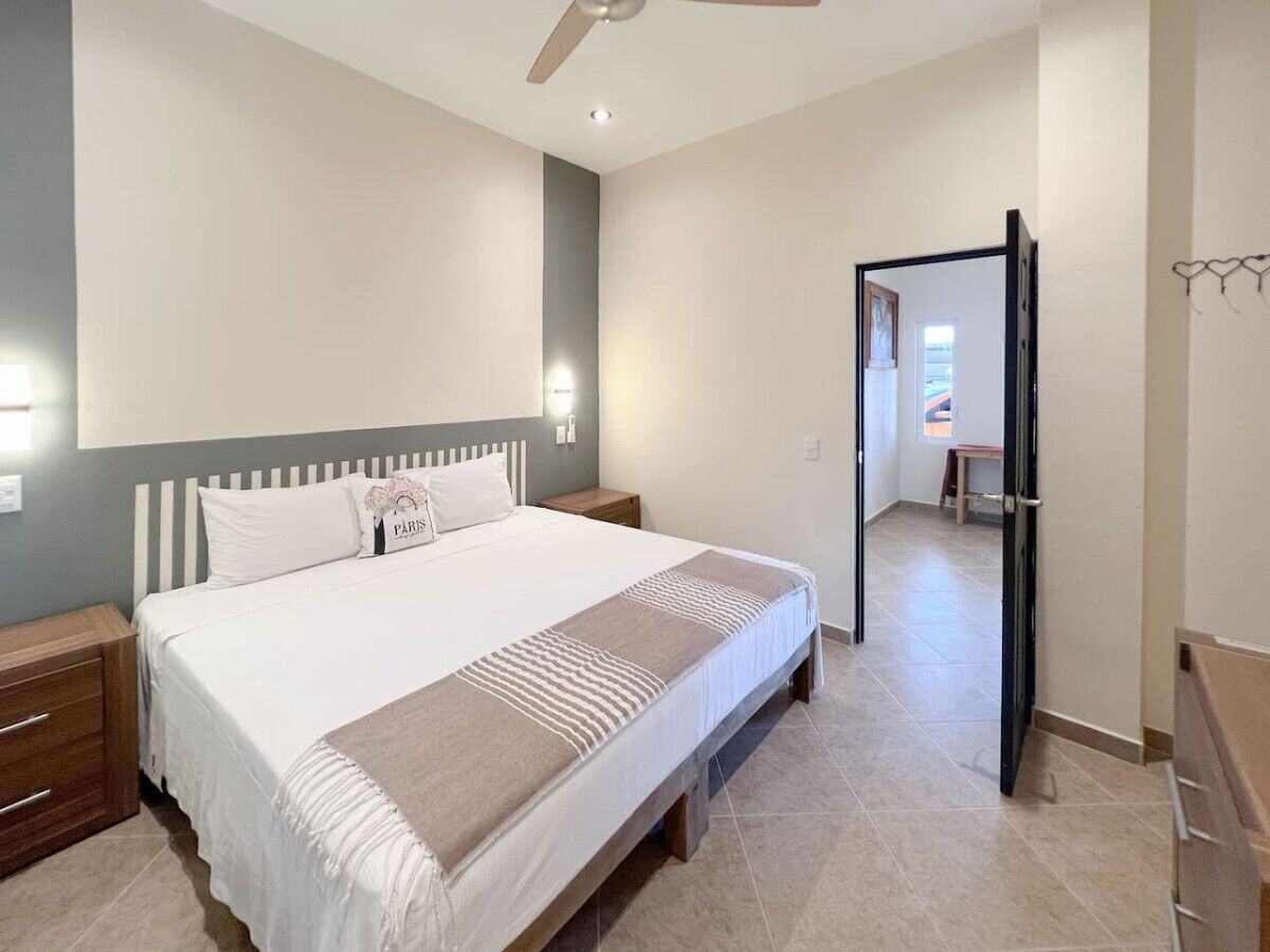 Un dormitorio con cama, mesitas de noche y un ventilador de techo, además de una puerta que da a otra habitación.