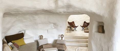 A cozy, rustic interior with a bed, pillows, a woven bench, and two chairs in a cave-like setting.