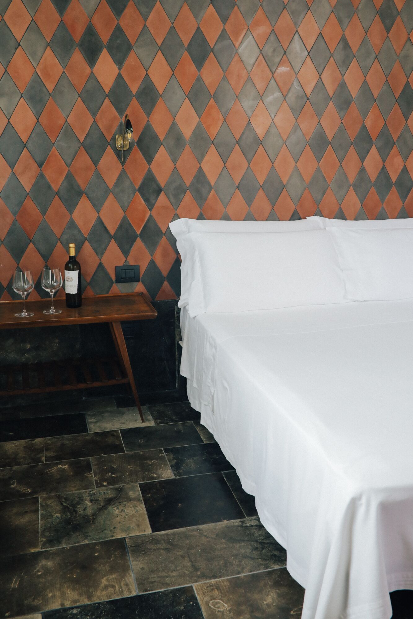 Une chambre d’hôtel avec un lit, une petite table sur laquelle se trouvent une bouteille et des verres, et un mur orné d’un motif.