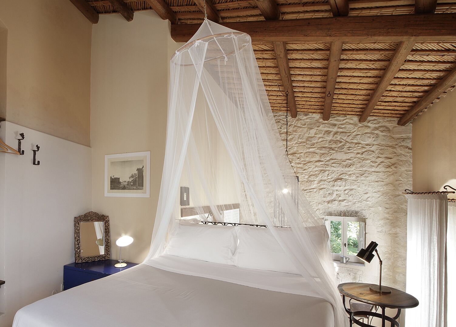 Een slaapkamer met hemelbed, een stenen muur, een klein tafeltje met lamp en een spiegel op een dressoir.