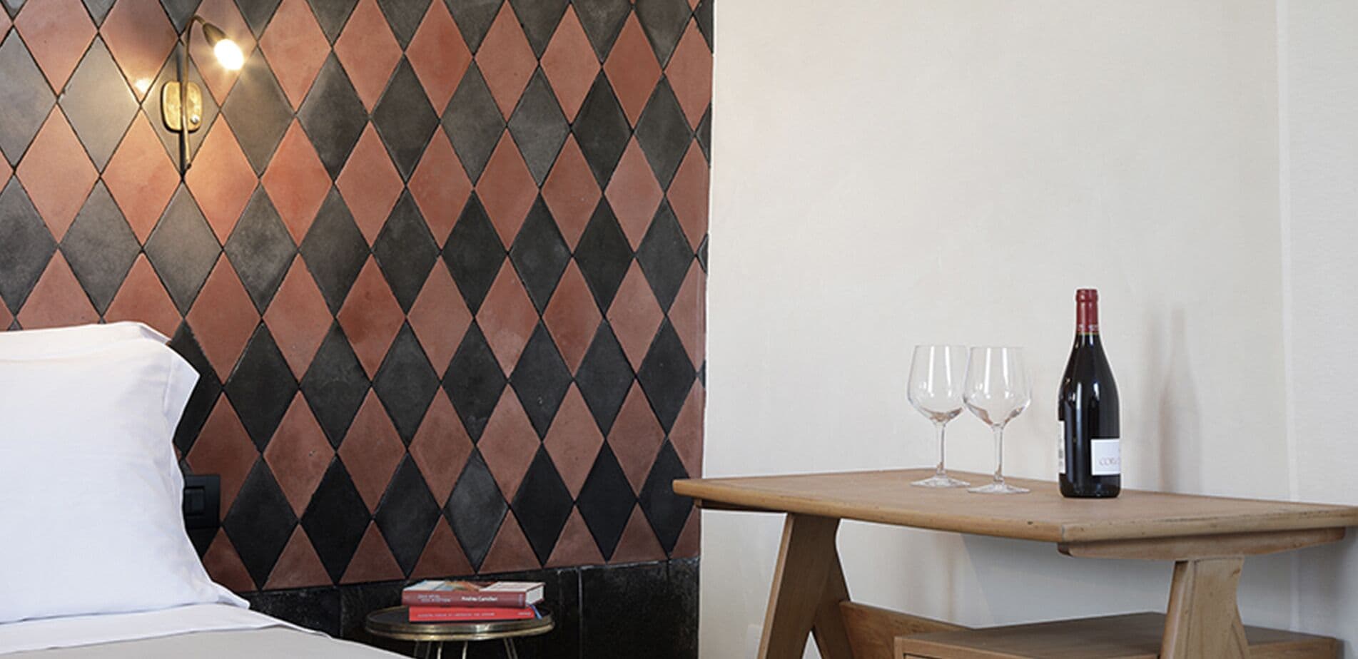 Quarto de hotel com uma cama, uma pequena mesa com uma garrafa de vinho e duas taças, e uma parede com design de azulejos em padrão losango.