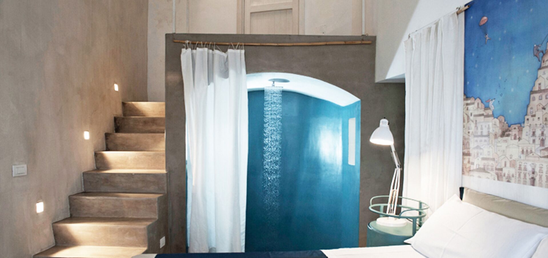 A bedroom with a staircase leading to a bathroom, a bed, a nightstand, a lamp, and a cityscape mural.