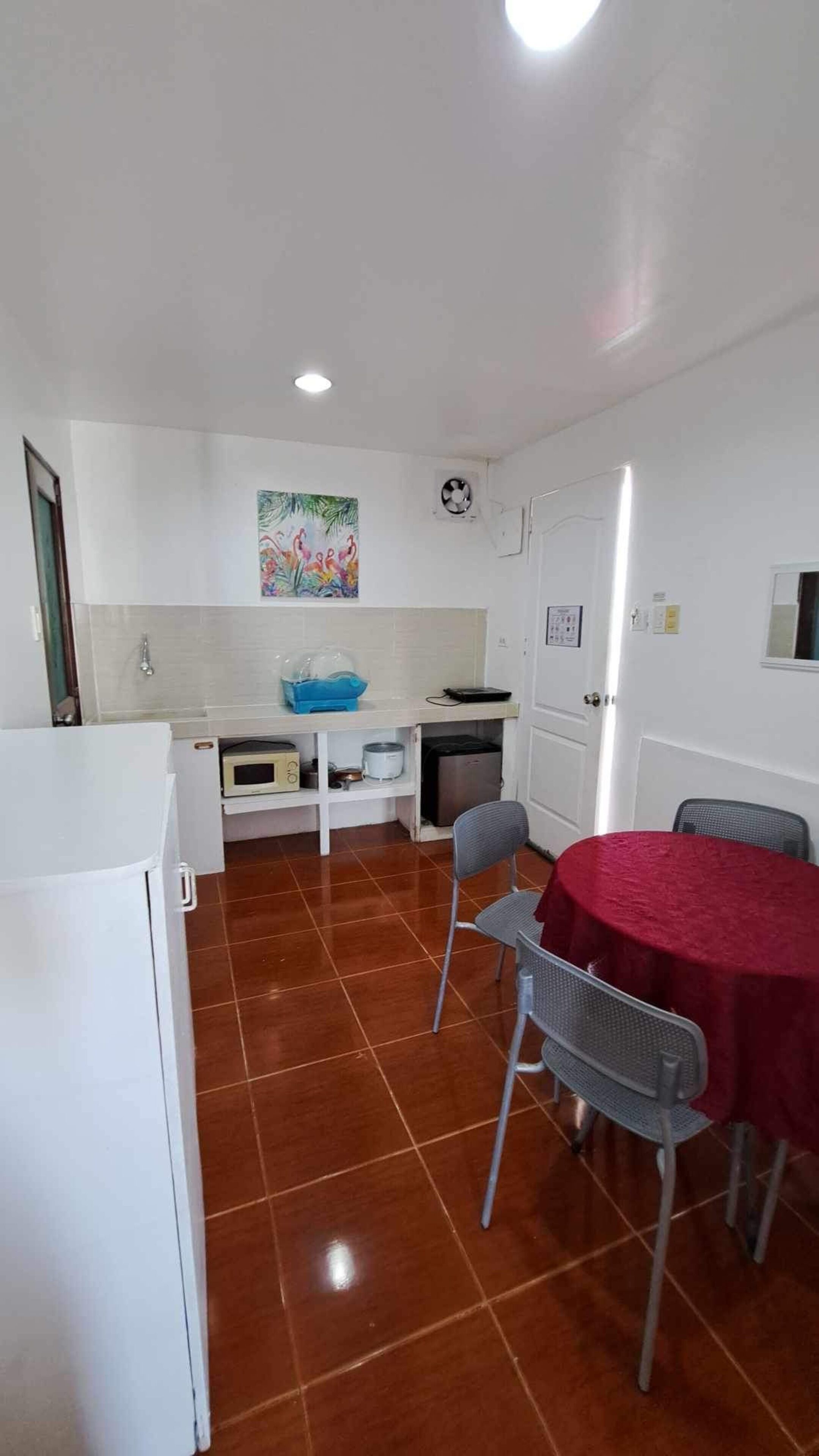 Cocina compacta con microondas, lavamanos y un refrigerador pequeño. Hay una mesa de comedor con mantel rojo y dos sillas. Un cuadro de colores está en la pared sobre el lavamanos.