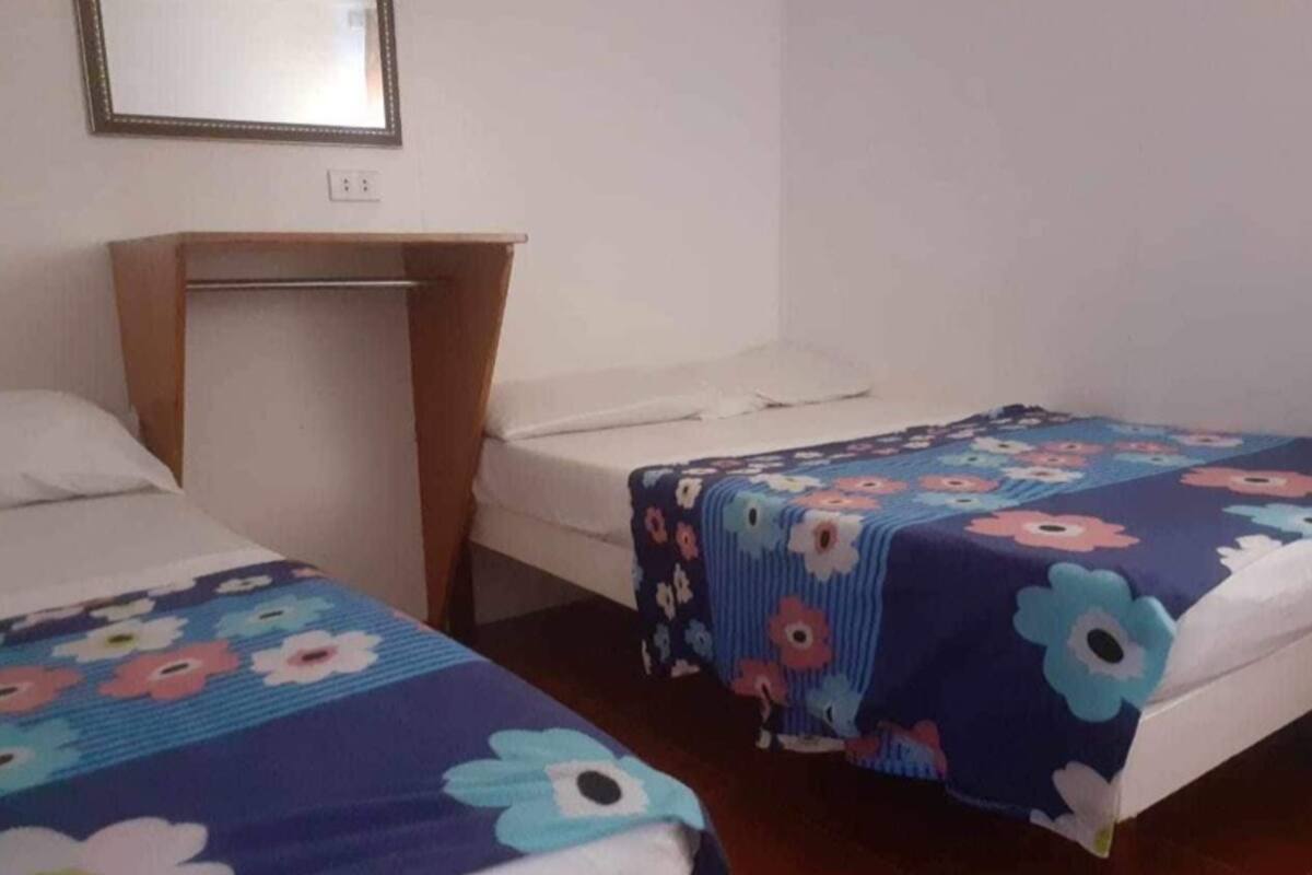 A room with two beds, each with a floral bedspread, a wooden bench, and a white wall with a framed mirror.