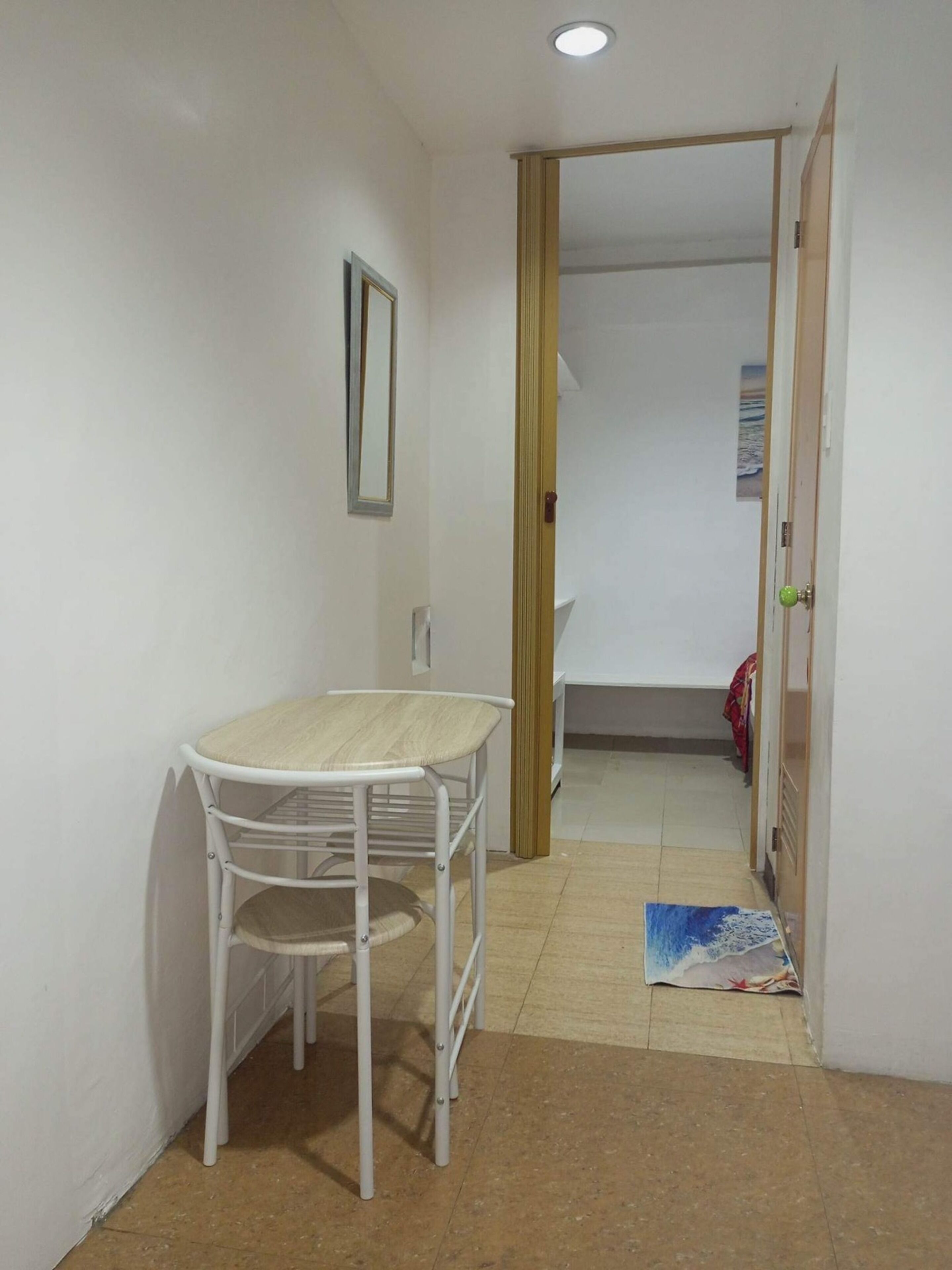 Una habitación pequeña y limpia con una mesa y una silla blancos, una puerta de madera y piso de baldosas.