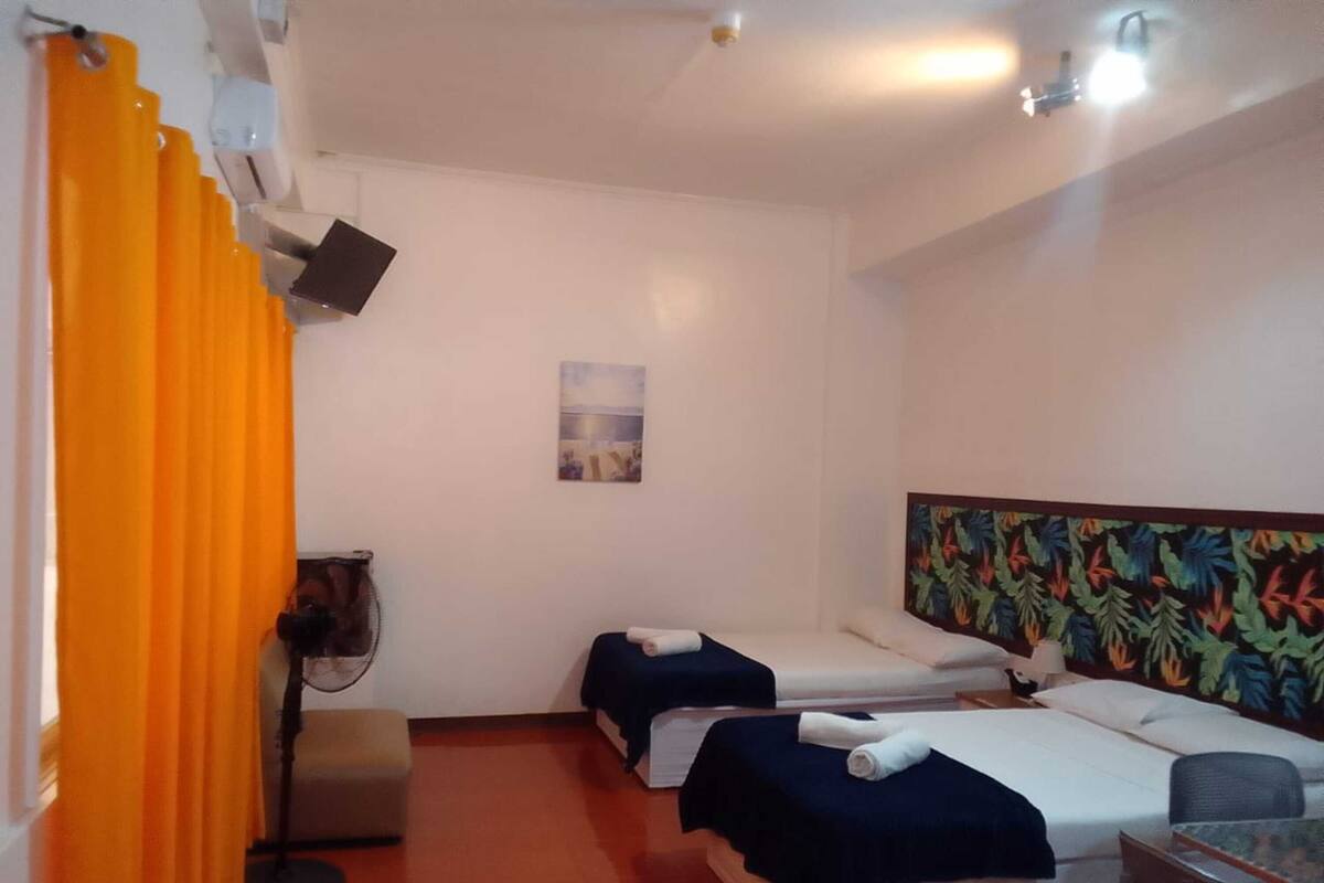 A room with two beds, a small table, a chair, a fan, and a painting on the wall.