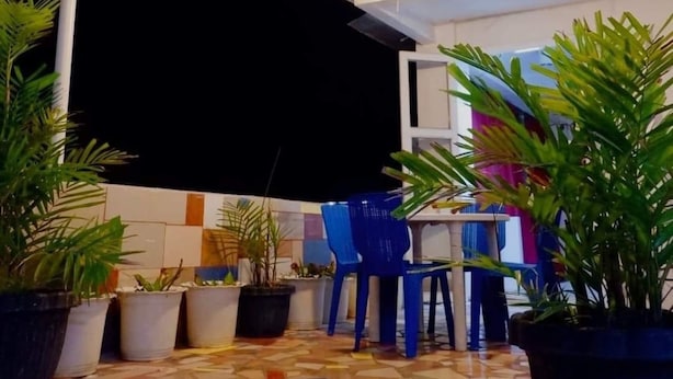 Uma área de estar ao ar livre com piso ladrilhado, vasos de plantas, cadeiras azuis e luzes de cordão.