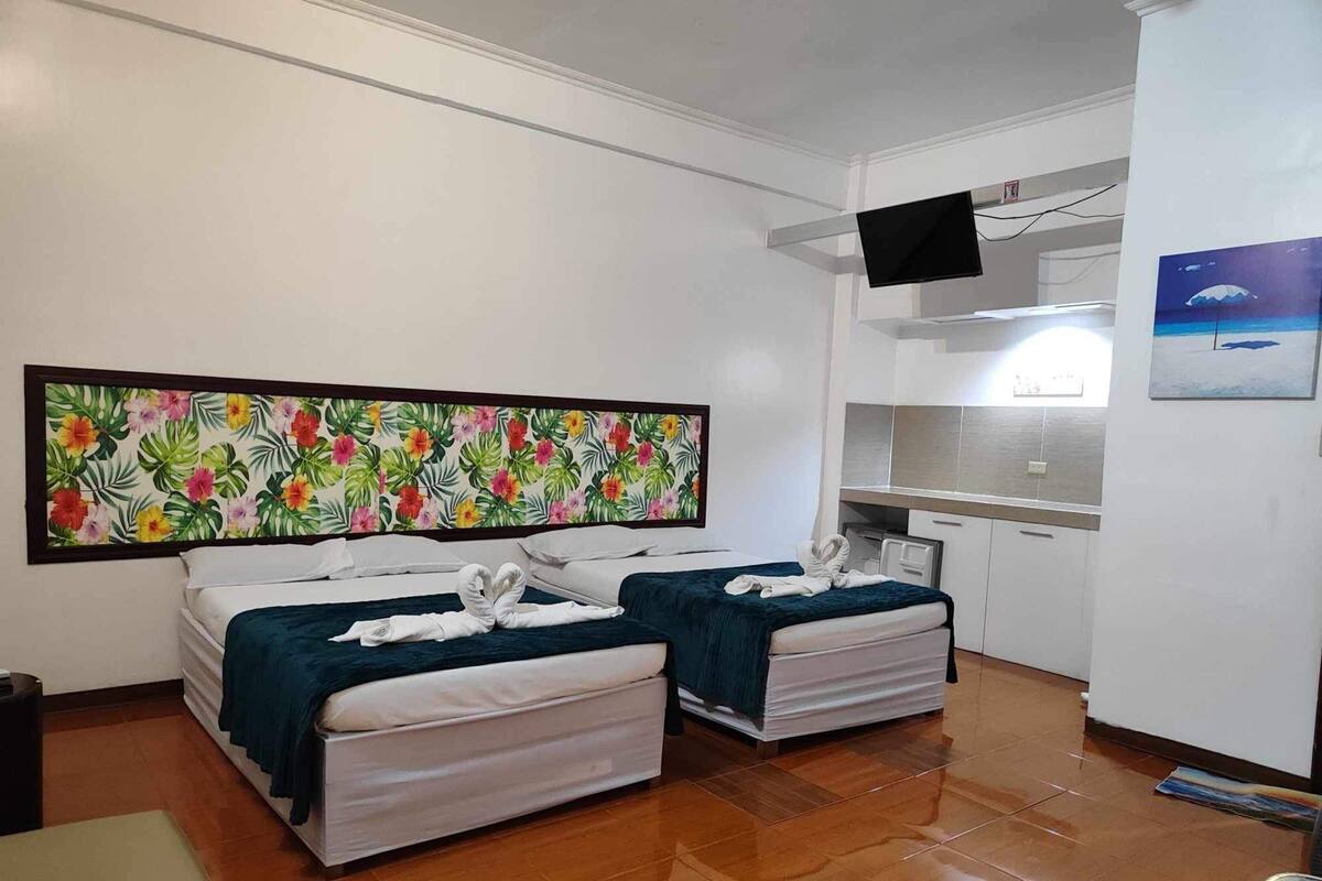 A hotel room with two beds, a large floral artwork on the wall, a small kitchen area, and a mounted TV.
