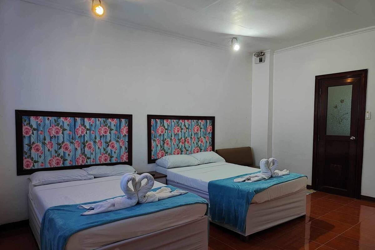 A hotel room with two single beds, each with a blue blanket and a heart-shaped towel on the bed. The room has white walls and a wooden door with a frosted glass panel.