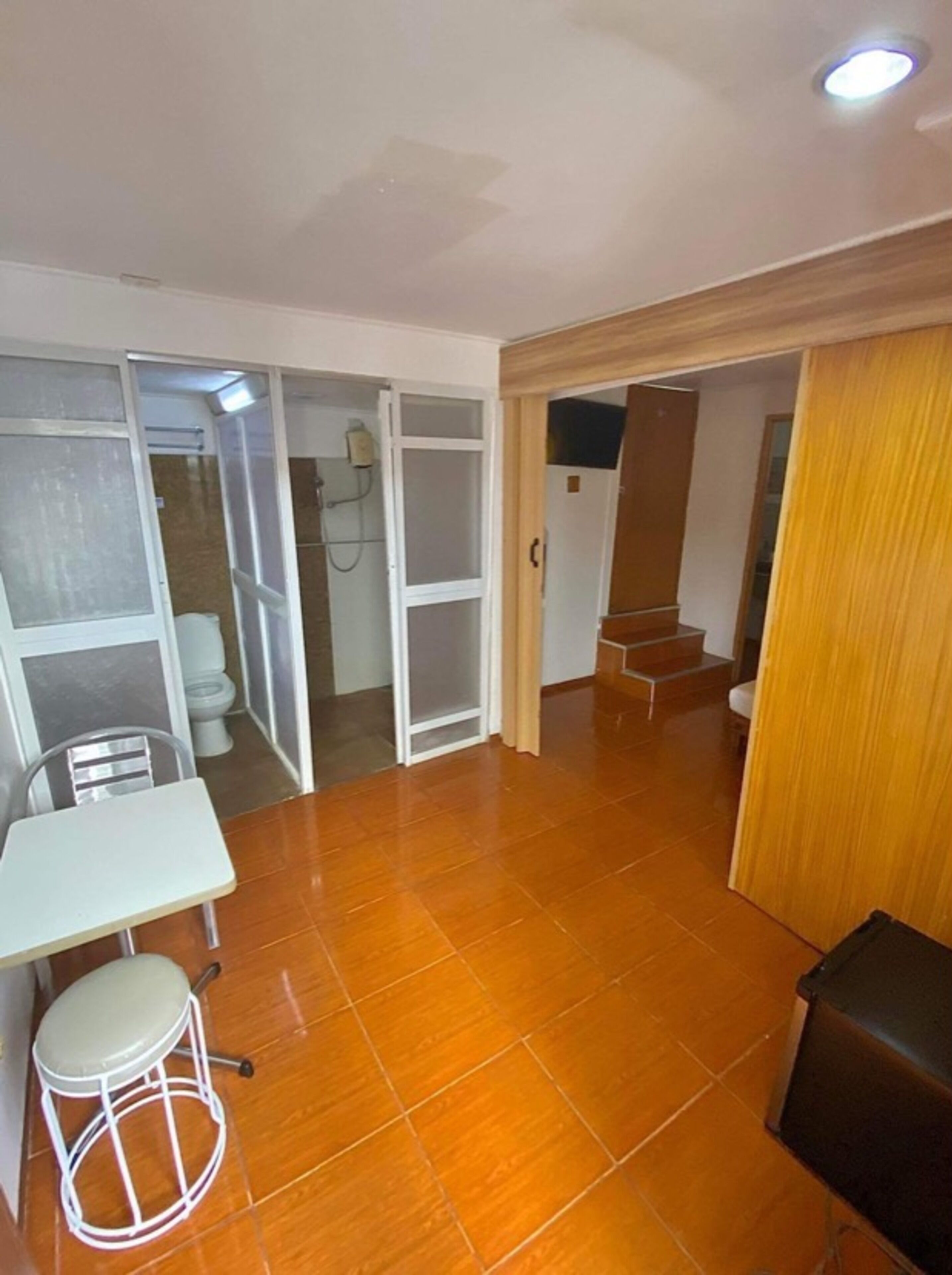 A room with a tiled floor, a white table with a stool, a shower area with a glass door, and a staircase leading to another level.