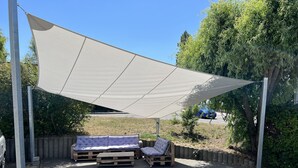 Un espacio al aire libre con una gran vela de sombra, dos bancos acolchados y una mesa de madera.