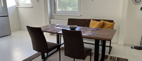 Un coin repas avec une table en bois, deux chaises, un coussin jaune et une fenêtre donnant sur des arbres.