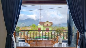 A balcony with wicker chairs and a table, overlooking a green house with a red door and a small garden.