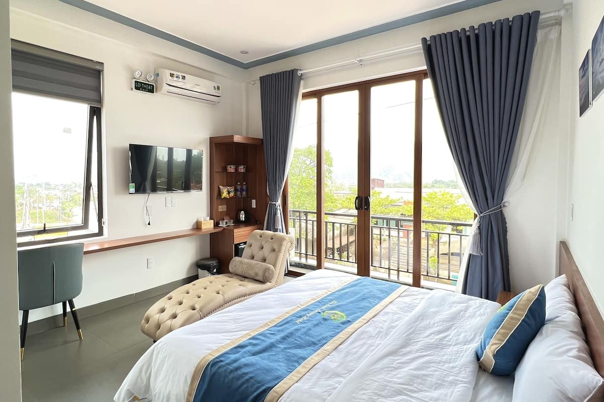 A hotel room with a large bed, a desk, a TV, and a balcony with curtains.