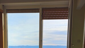 A room with a large sliding glass door leading to a balcony with a view of the sea and distant mountains. There are two potted plants on the floor. - Holiday Home Sunrise (Omis)