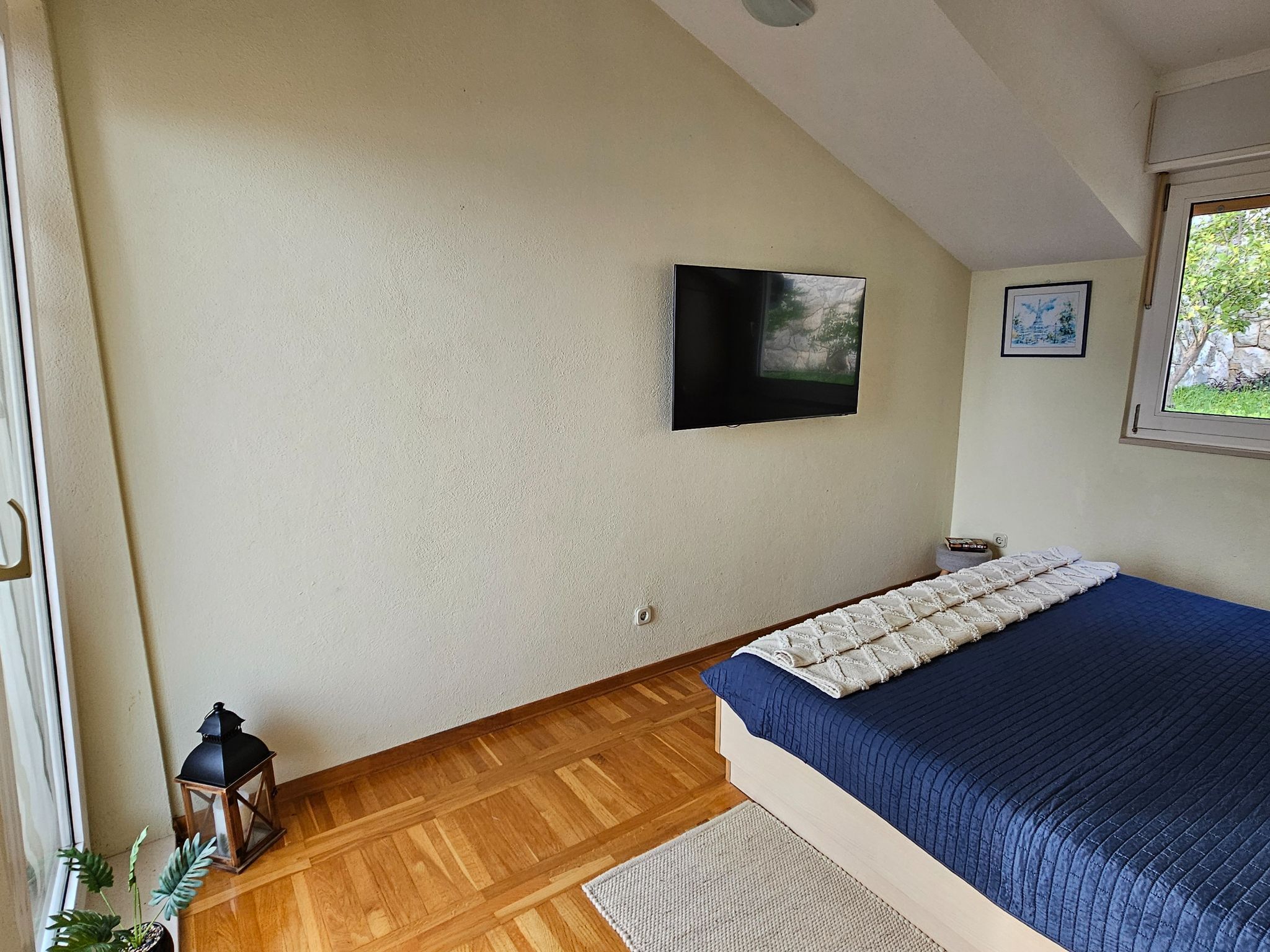 Ein Schlafzimmer mit einem Bett, einem an der Wand befestigten Fernseher, einem Fenster mit Blick auf Grünflächen und einem Holzboden.