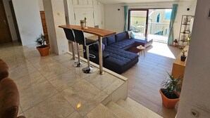 A modern living room with a dark blue sofa, a wooden dining table with two chairs, and a small round table with a single chair. There is a balcony with a railing and a view of the outside. - Holiday Home Sunrise (Omis)