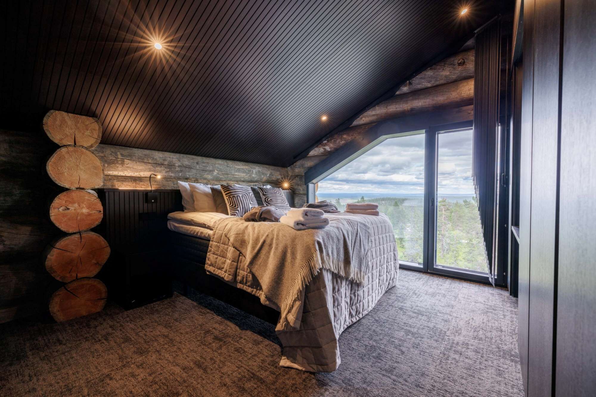 A bedroom with a large window, a bed with a blanket, and a wooden log wall.