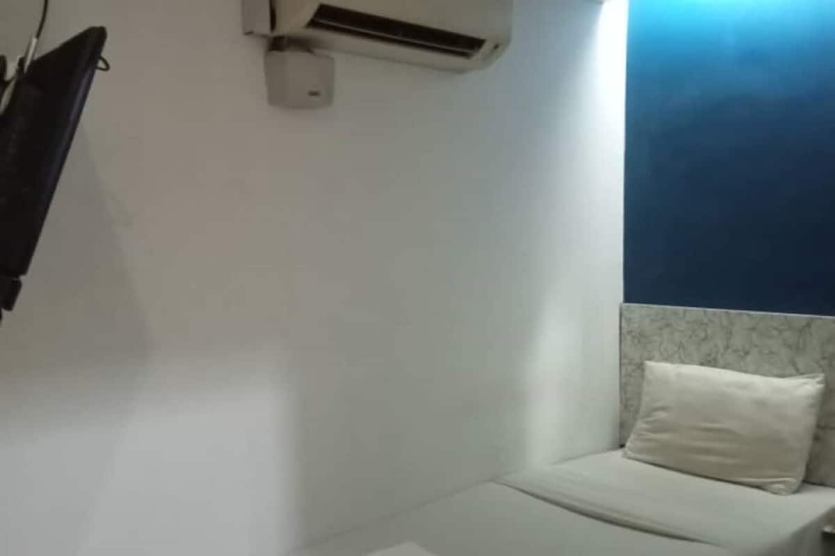 Ein kleines Zimmer mit einem Einzelbett, einer weißen Matratze, einem weißen Kopfkissen und einer weißen Decke. Über dem Bett ist eine Wand-Klimaanlage angebracht.