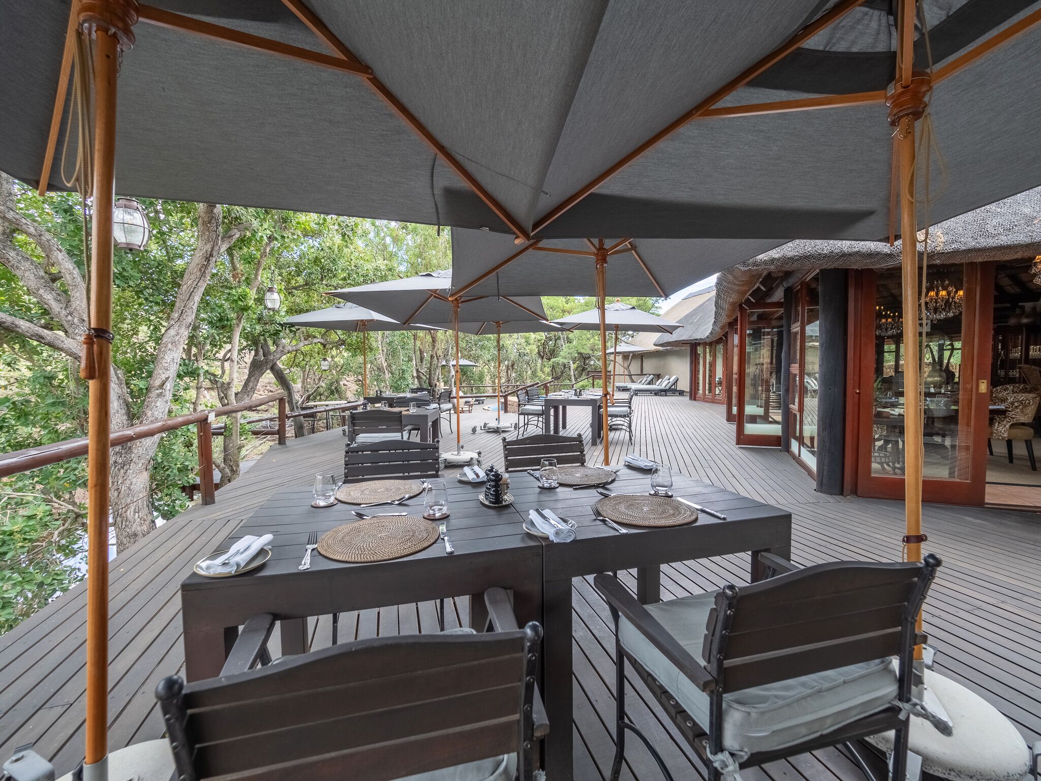 An outdoor dining area with tables and chairs, umbrellas, and a wooden deck overlooking a forested area.