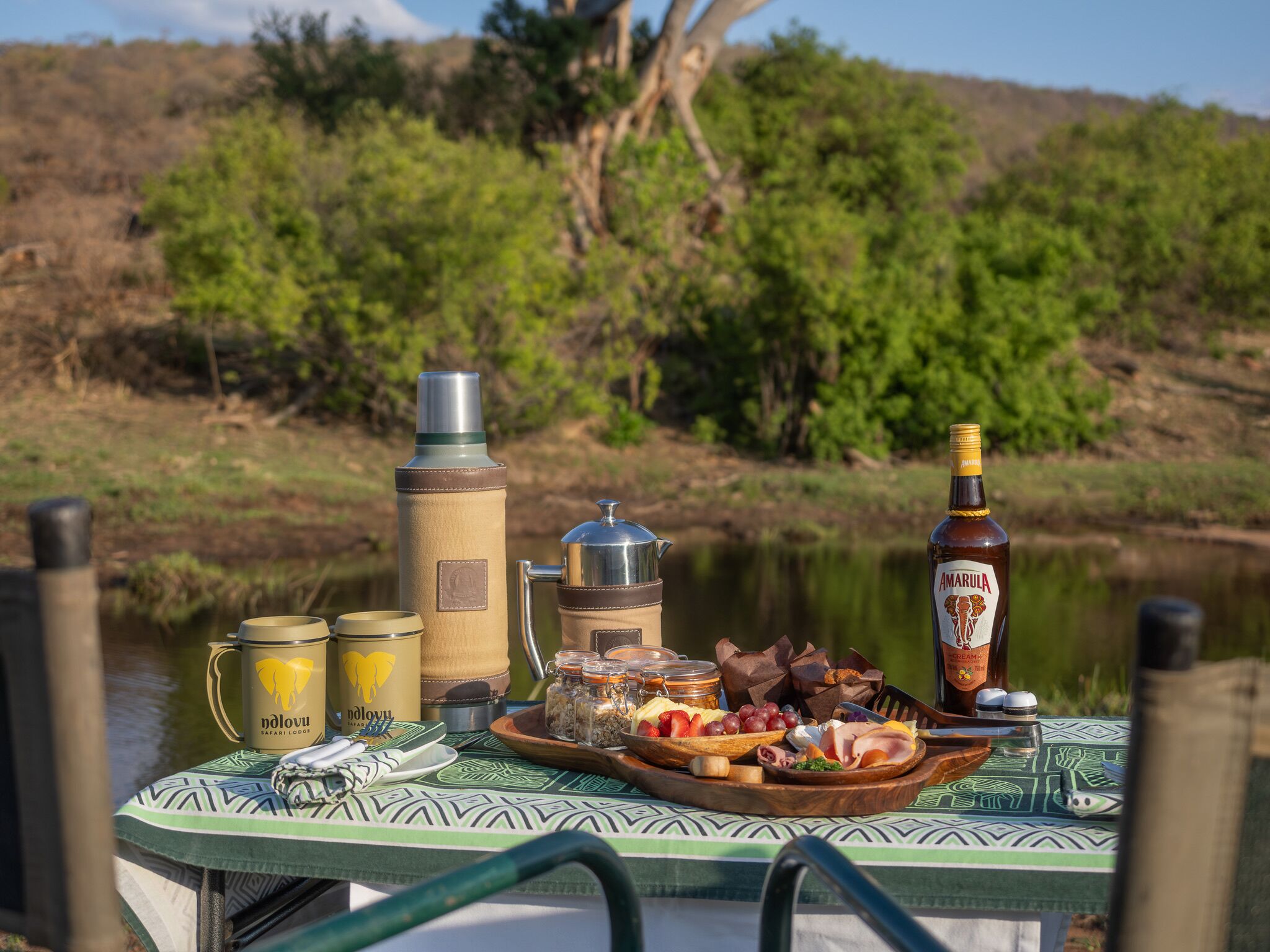 Una mesa dispuesta con diversos alimentos y bebidas, incluyendo una botella de Amarula, un termo y una taza, sobre una mesa con mantel de tela verde estampado.