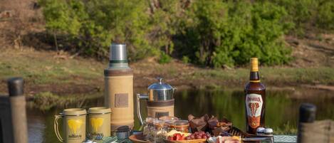 Una mesa dispuesta con diversos alimentos y bebidas, incluyendo una botella de Amarula, un termo y una taza, sobre una mesa con mantel de tela verde estampado.
