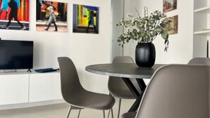 Un'area pranzo moderna con un tavolo rotondo, sedie grigie e un vaso di fiori. Alle pareti si trovano dipinti astratti e una TV a schermo piatto su un mobile bianco.